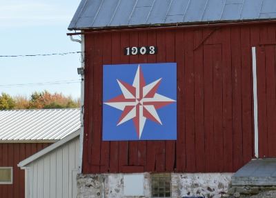 Red and white compass star on a barn quilt with a blue background on a red barn. 