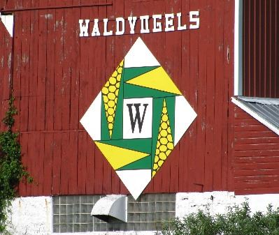 Green, yellow, and white barn quilt in a diamond pattern on a red barn. The barn quilt has a large &
