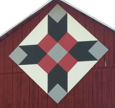 Barn quilt in a diamond pattern on the side of a red barn. The bar quilt has a white border with a b