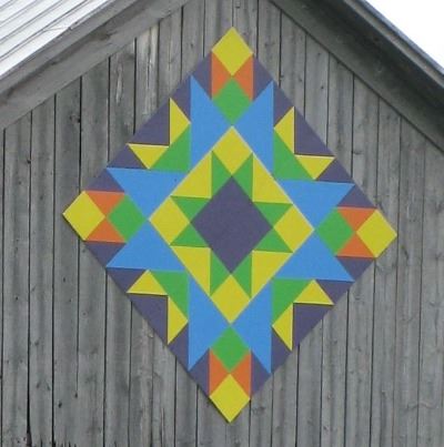 Barn quilt in a diamond pattern on the side of an unpainted barn. Pattern is a multicolored star bur