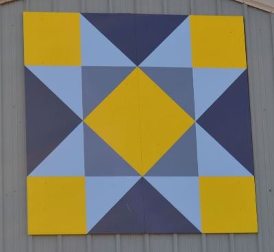 Barn quilt of a blue and yellow star in a square shape on a gray colored barn. 