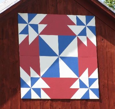 Barn quilt on the side of a red barn. Pattern is blue and white triangles with a red background.