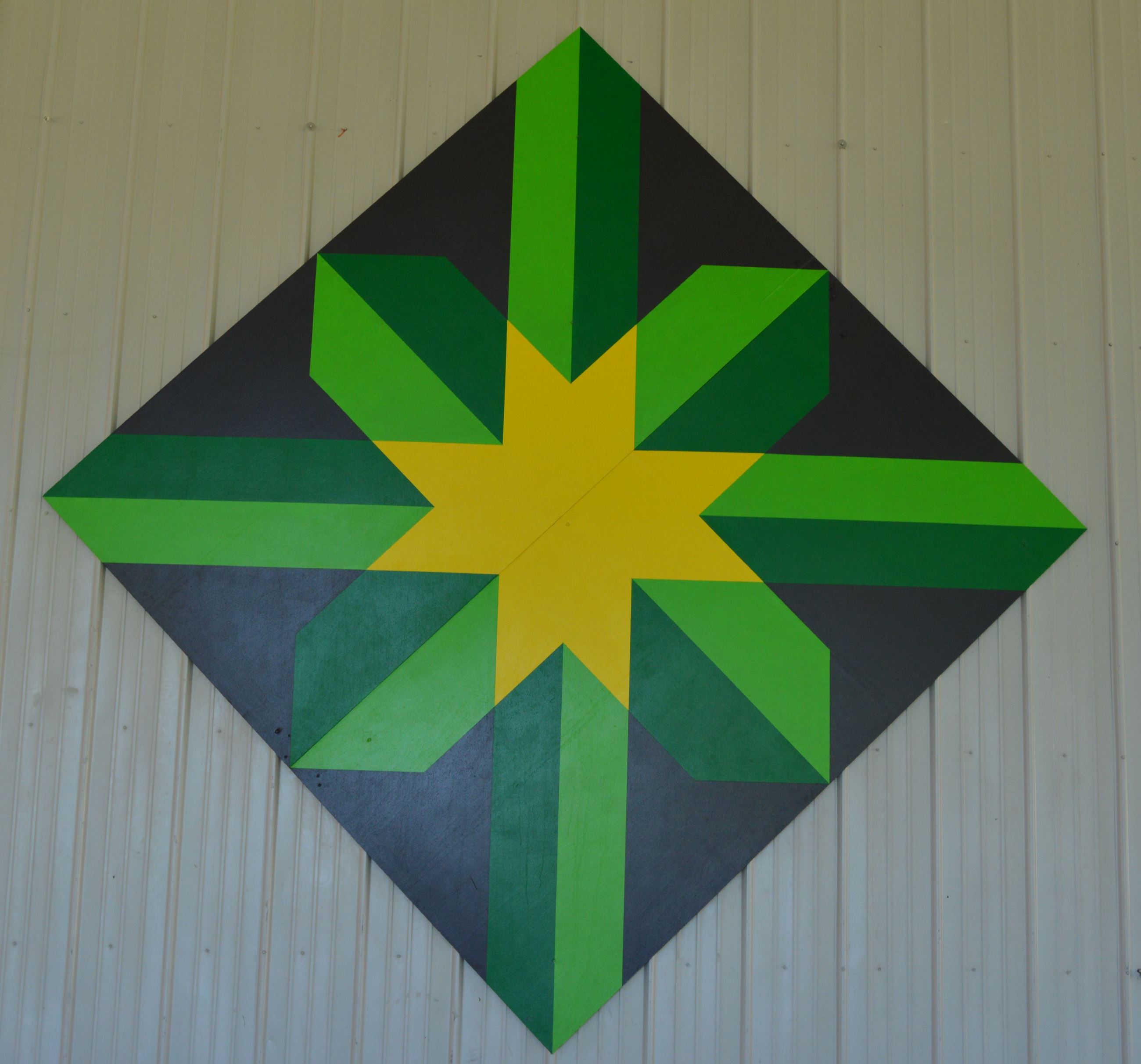 Barn quilt in a diamond shape on the side of a white steel wall. Pattern includes green intersecting