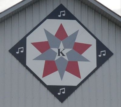 Barn quilt in a diamond shape on the side of a red and white barn. Barn quilt pattern includes a gra