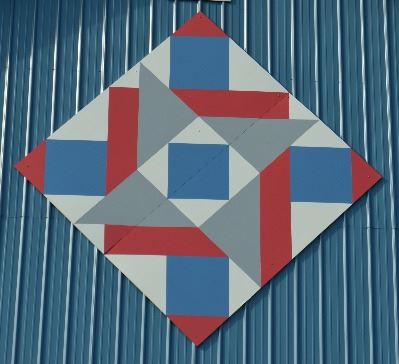 Barn quilt in a diamond shape on the side of a blue barn. Barn quilt pattern includes squares, trian