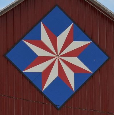 Barn quilt in the shape of a diamond on the side of a red barn. Barn quilt pattern in the shape of a