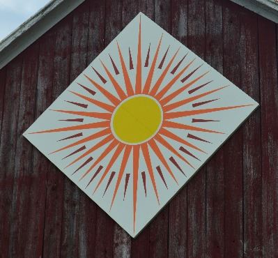 Barn quilt in a diamond shape on the side of a red barn. Pattern is of a sun in the center with red 