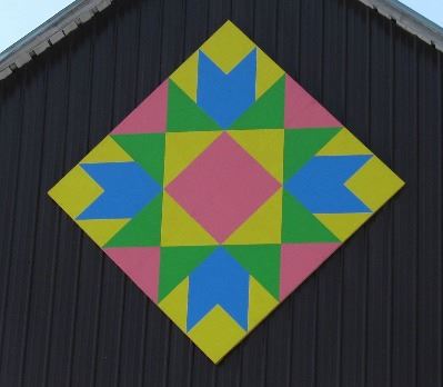 Barn quilt in a diamond pattern on the side of a brown and white barn. Pattern is of various triangl