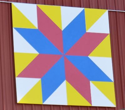 Barn quilt on the side of a red barn. Pattern is of a red and blue star burst with a white and yello