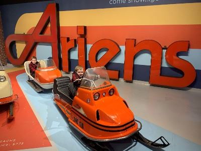 Two boys each on stationary, orange snowmobiles with a large Ariens sign behind them